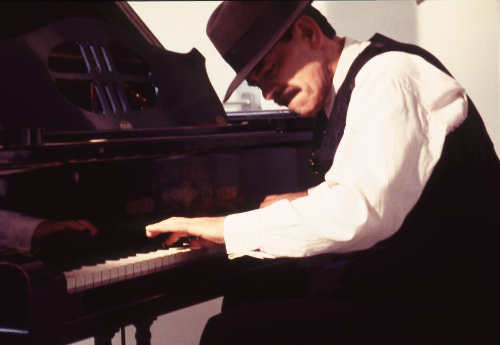 A color photo of John Larkin wearing a white shirt and a black waistcoat and fedora, playing a grand piano