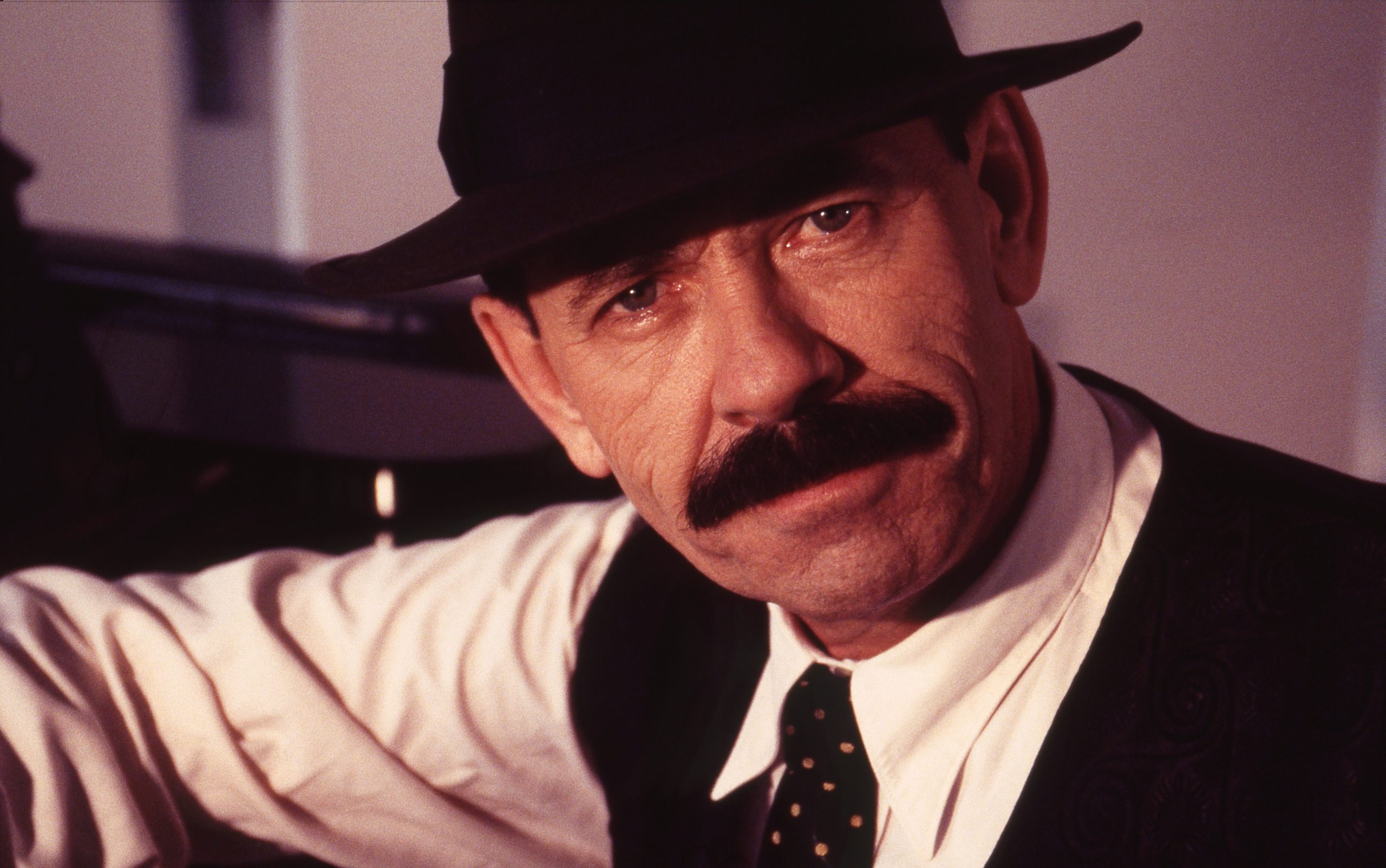 A close up portrait of Scatman John, a middle aged man with a black moustache, who is wearing a fedora, shirt, waistcoat and tie. 