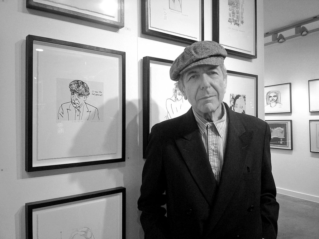 A black and white photo of Leonard Cohen, an older man in a flat cap in a suit and shirt, standing against drawings framed in a gallery.